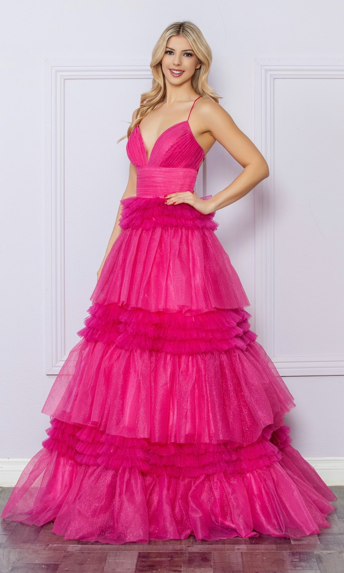 A woman in the Nox Anabel Ruffled Glitter Prom Dress R1316, featuring bright pink tiers and spaghetti straps, stands smiling in front of a light-colored wall.
